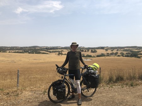 Cyclist Theo Foster stands in front of his bike wearing a hat, long sleeve t-shirt, cycling shorts and shoes. HIs bike is carrying four bags. A yellow wheat field is in the background.