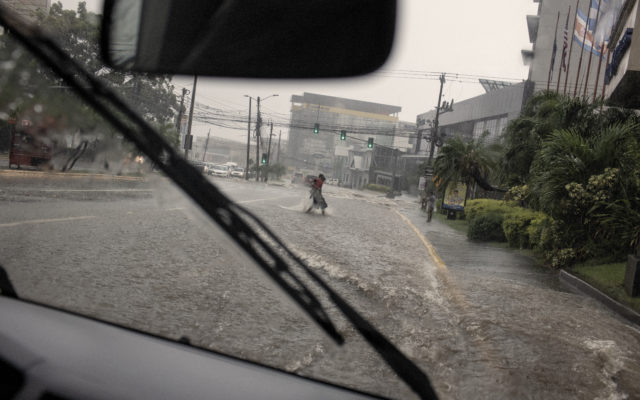 The view from a car window. A person crosses a flooded street during a storm carrying a child.